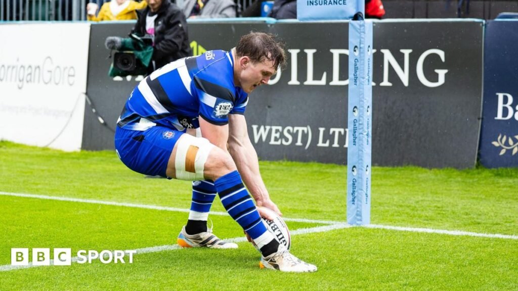 Ted Hill, wearing a blue shirt with black and white stripes, placing the ball over the try line to score near the corner flag during a game between Bath and Newcastle.