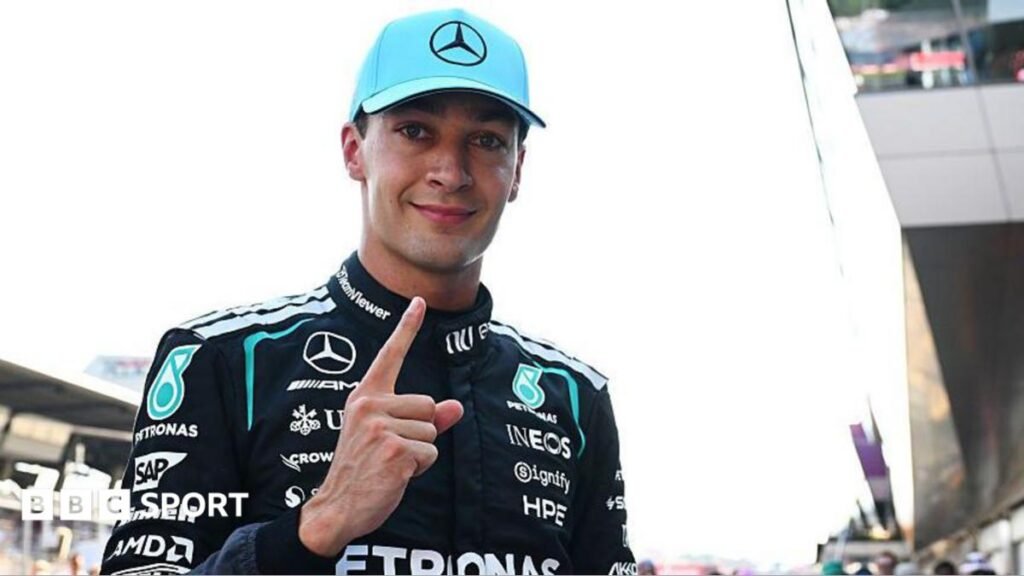 George Russell looks at the camera and points his index figure upwards