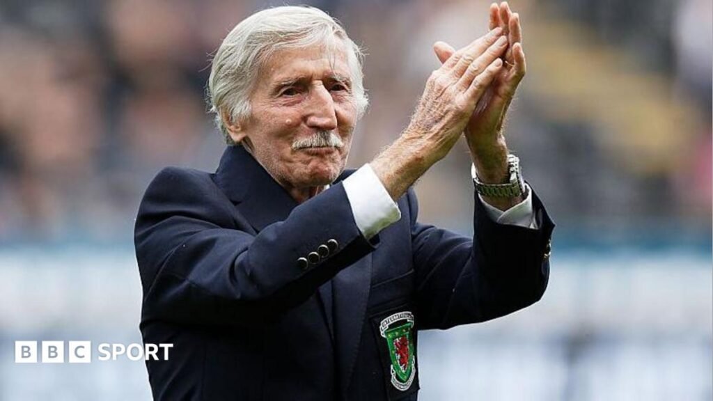 Cliff Jones: Wales and Tottenham Hotspur legend made MBE Cliff Jones, who has white short hair and a moustache, has his arms raised clapping. He has a blue blazer on with the Wales logo.