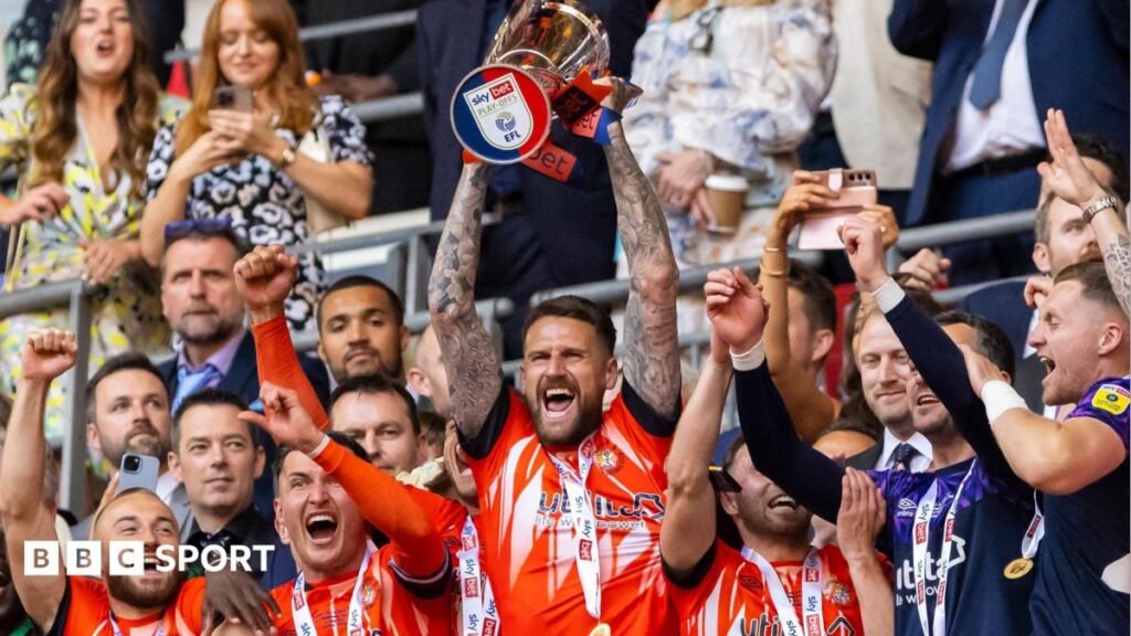 Six of the Luton Town team are visible on Wembley balcony holding EFL Trophy, wearing either orange home kit, or training tops