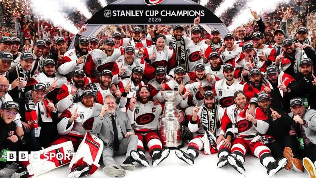 The Carolina Hurricanes players, wearing white and red kits, gathered around the silver Stanley Cup trophy