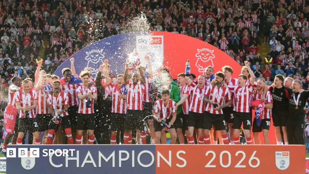 Lincoln City's players, wearing their red and white striped home shirt with black shorts and red socks, lift the League One trophy. Champagne can be seen getting sprayed into the air. The fans can be seen behind the stand which says "CHAMPIONS 2026" on it.
