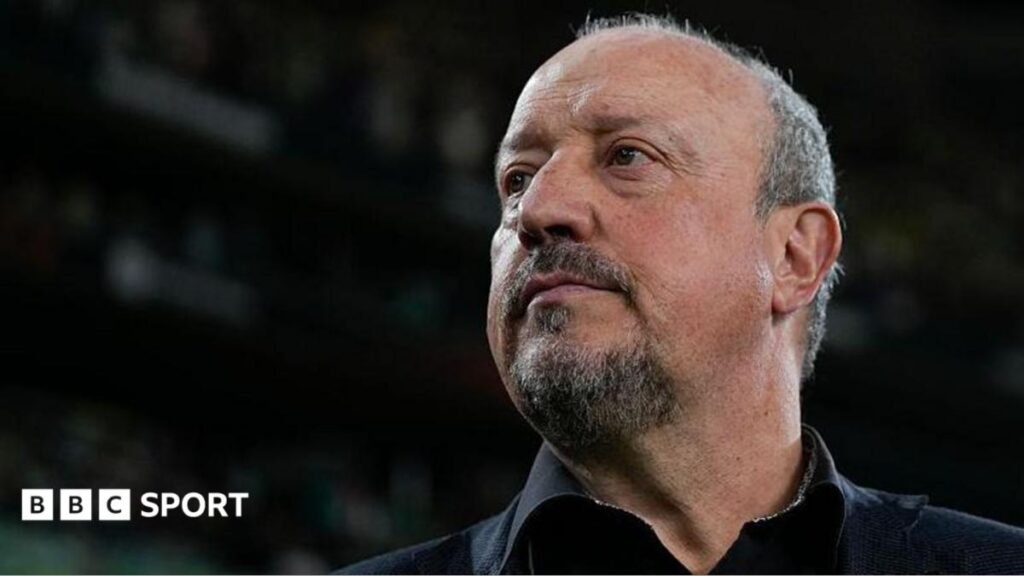 Rafa Benitez open to Scotland job – what would he bring and is he affordable? Rafael Benitez, wearing a suit and tie, holds his phone with two hands while watching a match