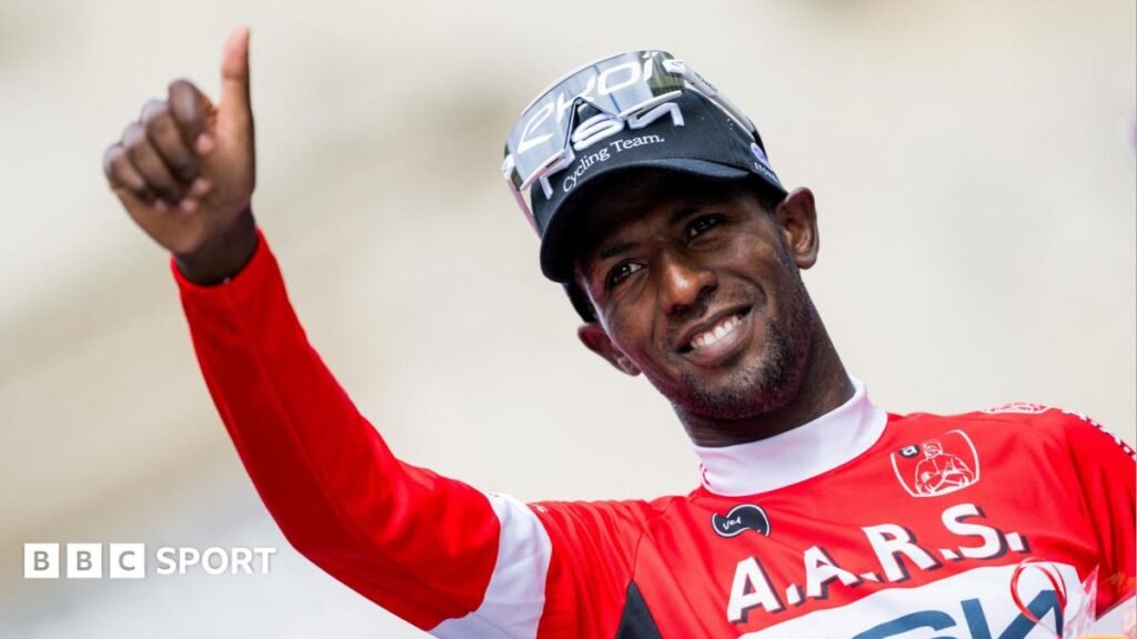 Tour de France 2026: Biniam Girmay aims to reclaim green jersey with NSN Cycling Biniam Girmay, visible from stomach up wearing a long-sleeved green cycling jersey, white cap and sunglasses pushed up on to the top of his head, holds both arms up as he smiles in celebration. He is on a post-race podium, discernible due to the sponsors logos visible on the yellow and green backdrop