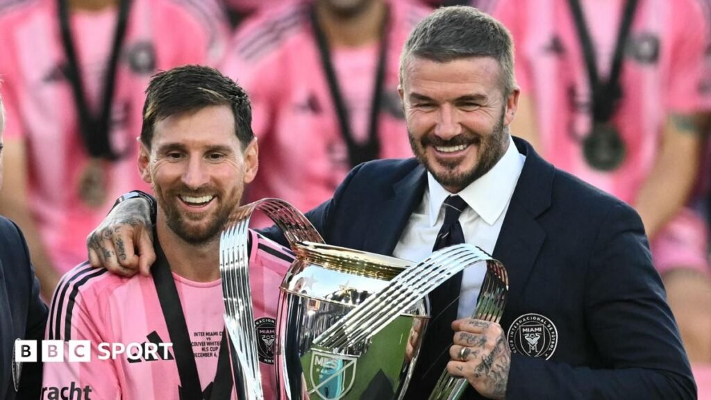 World Cup 2026: What next for Major League Soccer? Lionel Messi stands alongside David Beckham, who has his right arm around the Argentine as they show off the MLS Cup trophy. Messi is wearing Inter Miami's pink kit, while Beckham is dressed smartly in a club blazer, white shirt and dark tie. Both are smiling.