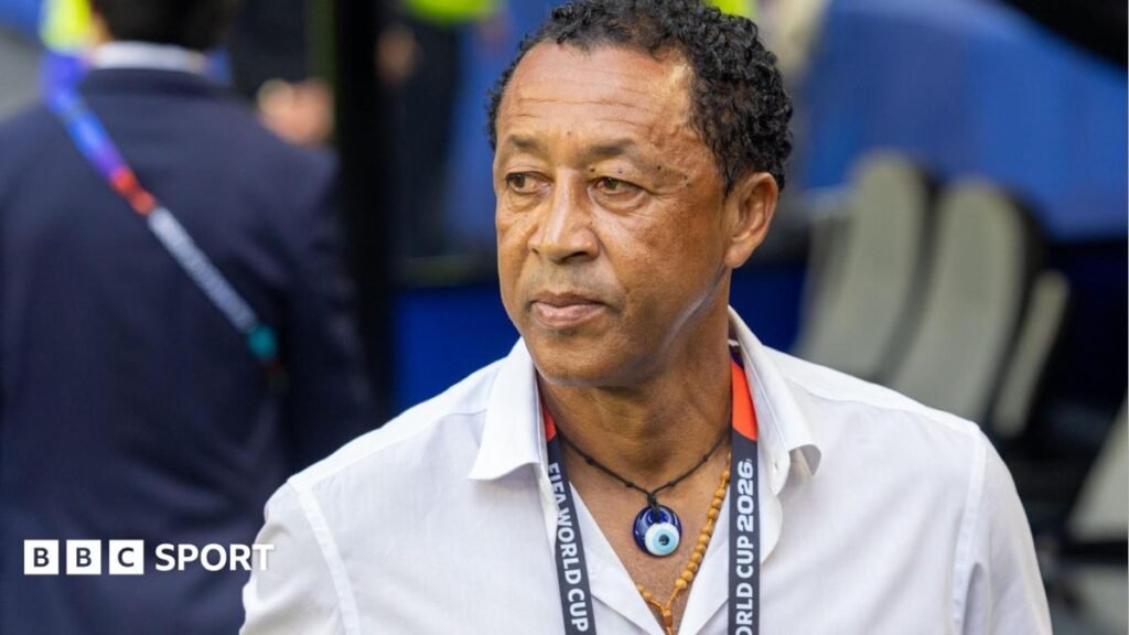 Bubista wears a white shirt, two necklaces and a World Cup 2026 lanyard around his neck as he looks to his right while standing in front of a dugout at at a football stadium