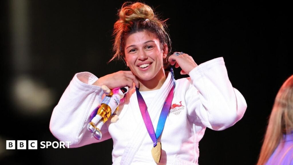Acelya Toprak, wearing England's white judo robe, smiles with her gold medal around her neck on the podium