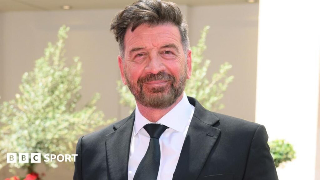 Nick Knowles looks on wearing a black suit and matching tie