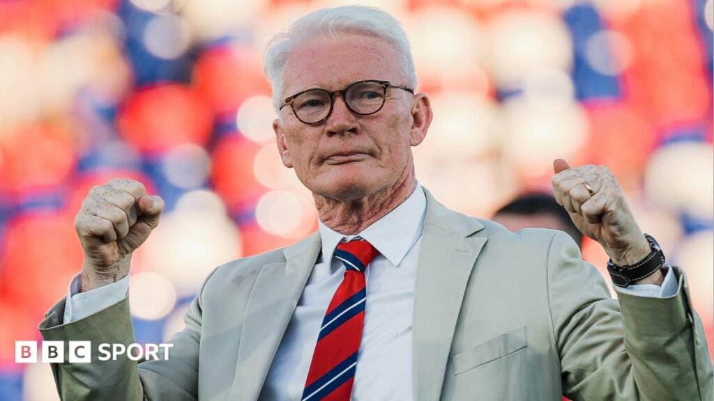 Eamonn McManus, wearing a beige suit jacket, light coloured shirt and red and blue tie, pumps both his fists in the air to celebrate after a St Helens game