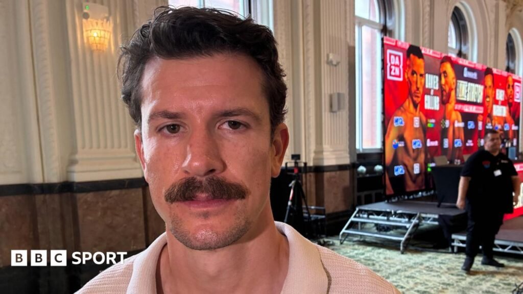 Stoke boxer Nathan Heaney to retire after Gerome Warburton rematch Boxer Nathan Heaney, who has dark hair and a moustache, stands to the side of a promotional dais advertising his next bout. A security man stands in the background to his left