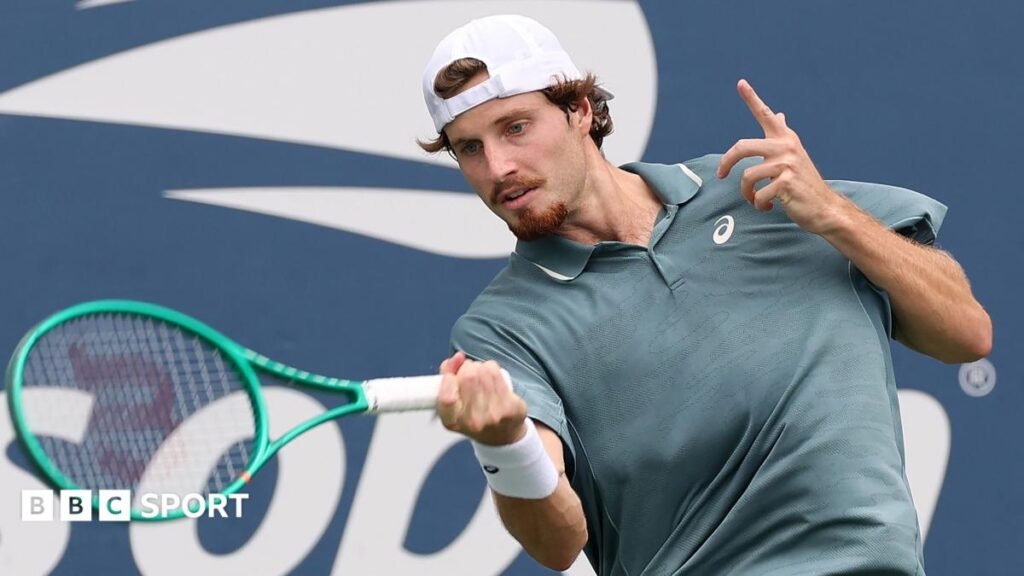 US Open 2026 results: Toby Samuel earns first Grand Slam main-draw win but Cameron Norrie suffers early exit Toby Samuel, wearing a grey shirt and white backwards baseball cap, hits the ball with a green racquet
