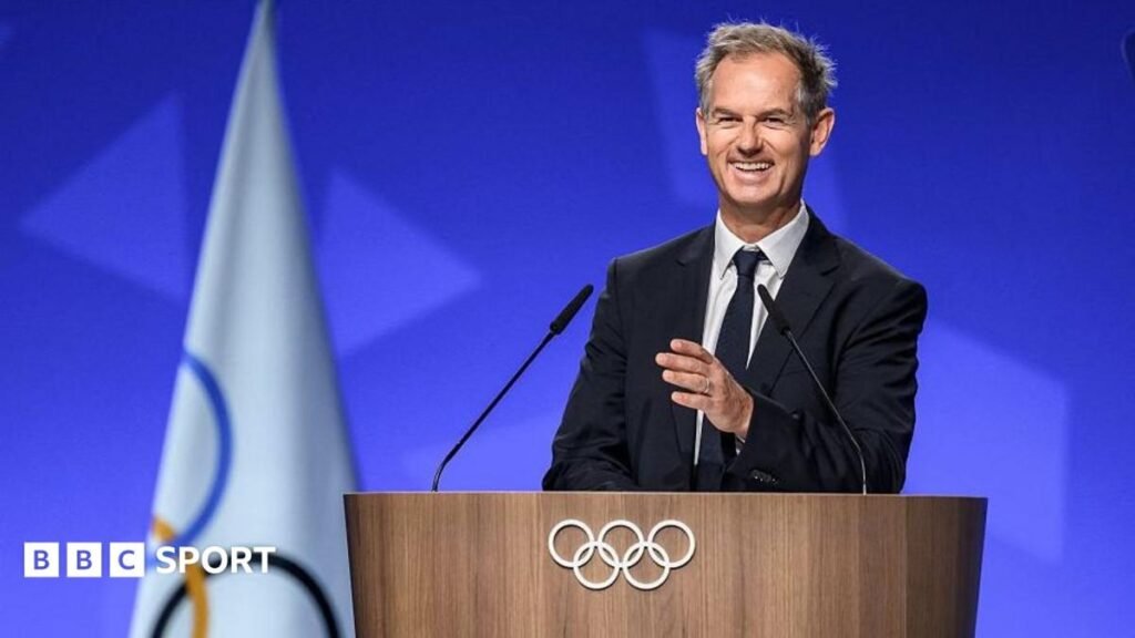 A smiling man in a dark suit and tie stands behind a wooden podium adorned with white Olympic rings with a folded white Olympic flag to his right