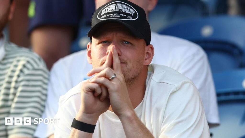 Aaron Ramsdale, wearing a black baseball cap and white t-shirt, watches Southampton's EFL Cup game at Colchester United from the stands