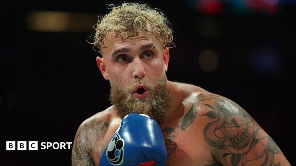Jake Paul holds up his hand and is wearing a blue boxing glove