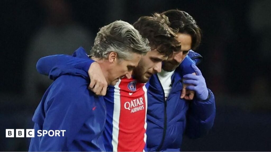 Khvicha Kvaratskhelia (centre, wearing a blue and red PSG shirt) is helped from the field by two men wearing dark blue jackets