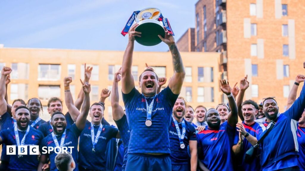 The captain of London Broncos lifts the Championship trophy as his team-mates lift their arms in celebration behind him