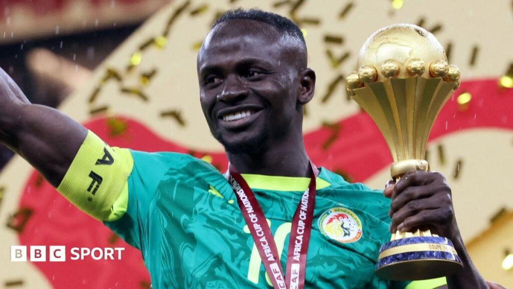 Sadio Mane, visible from chest up wearing a green Senegal shirt and with a yellow captain's armband on his right bicep, smiles as he holds the Africa Cup of Nations trophy in his left hand after the trophy presentation at Afcon 2025 in Morocco. He has a red ribbon around his neck, although the medal is out of shot, and golden ticker tape can be seen falling behind him